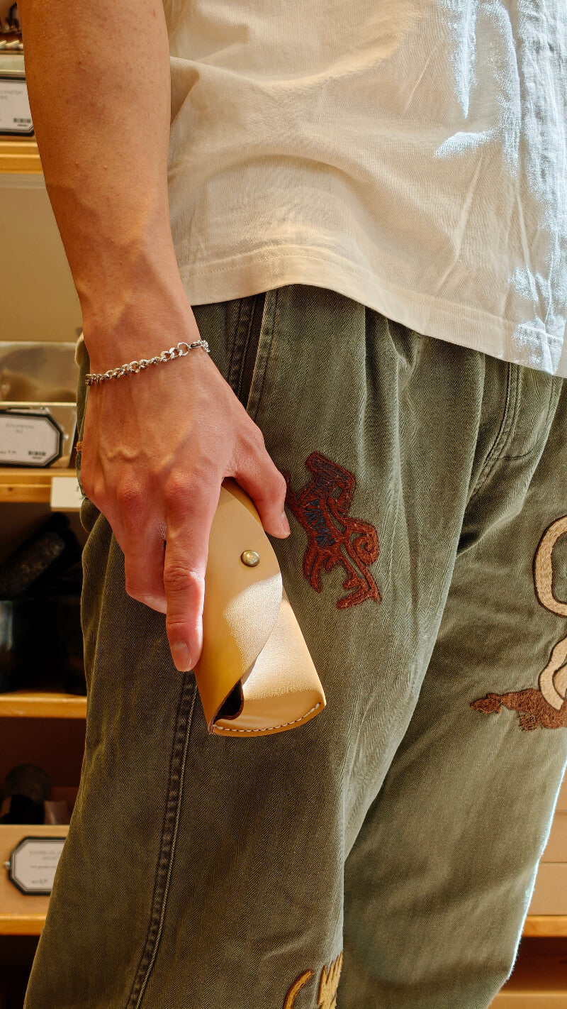 Person wearing green pants with embroidered designs, holding a small leather glasses case.